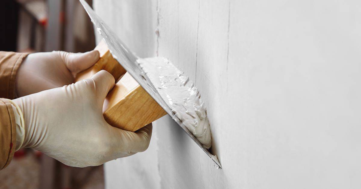 Hands wearing gloves applying white stucco with trowel on wall