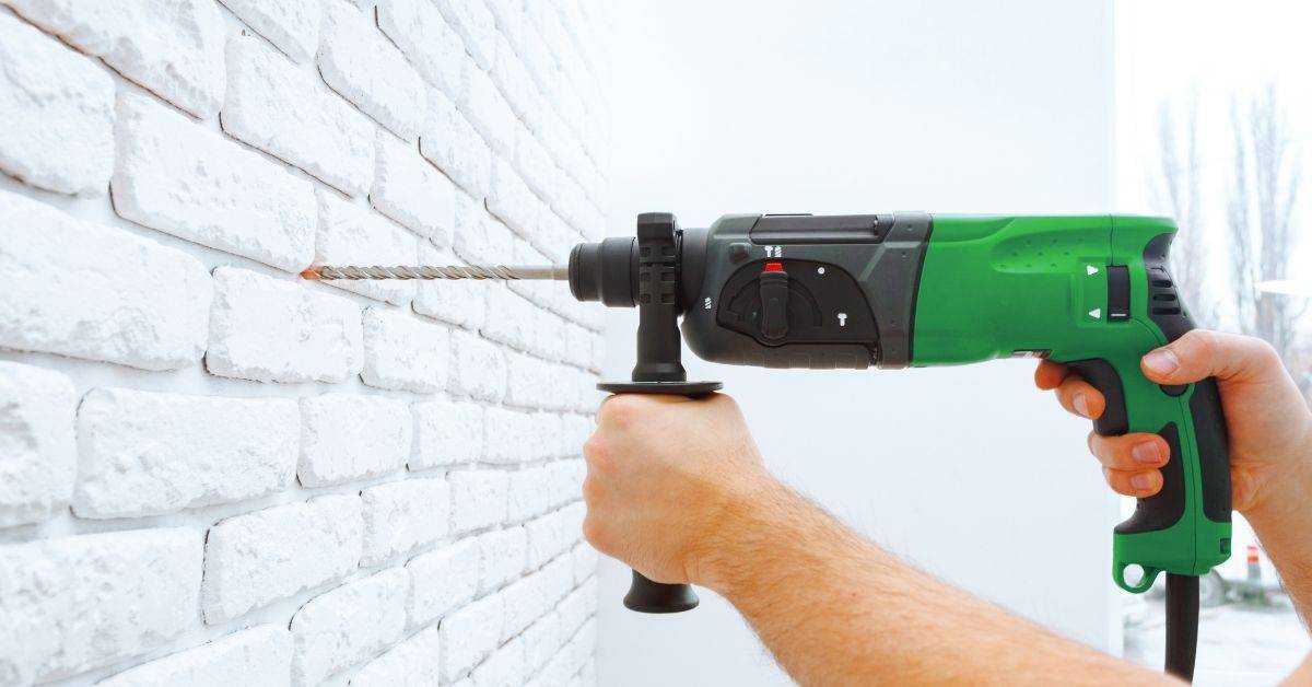 Hands holding green drill making hole in white textured brick wall