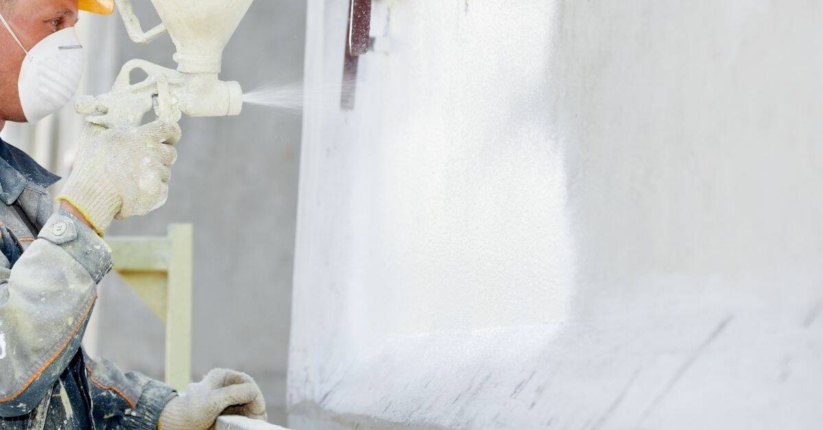 Person spraying white stucco sealer on wall wearing mask and gloves