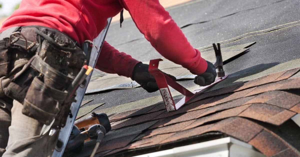 Person wearing red shirt installing shingles using hammer and nail