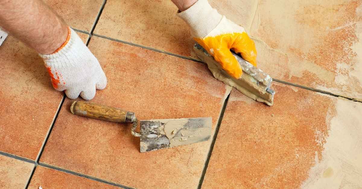 Hands applying grout between reddish-brown floor tiles with tools