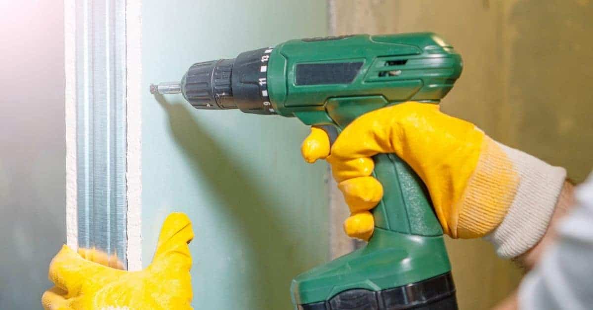 Hands wearing yellow gloves using green cordless drill on drywall