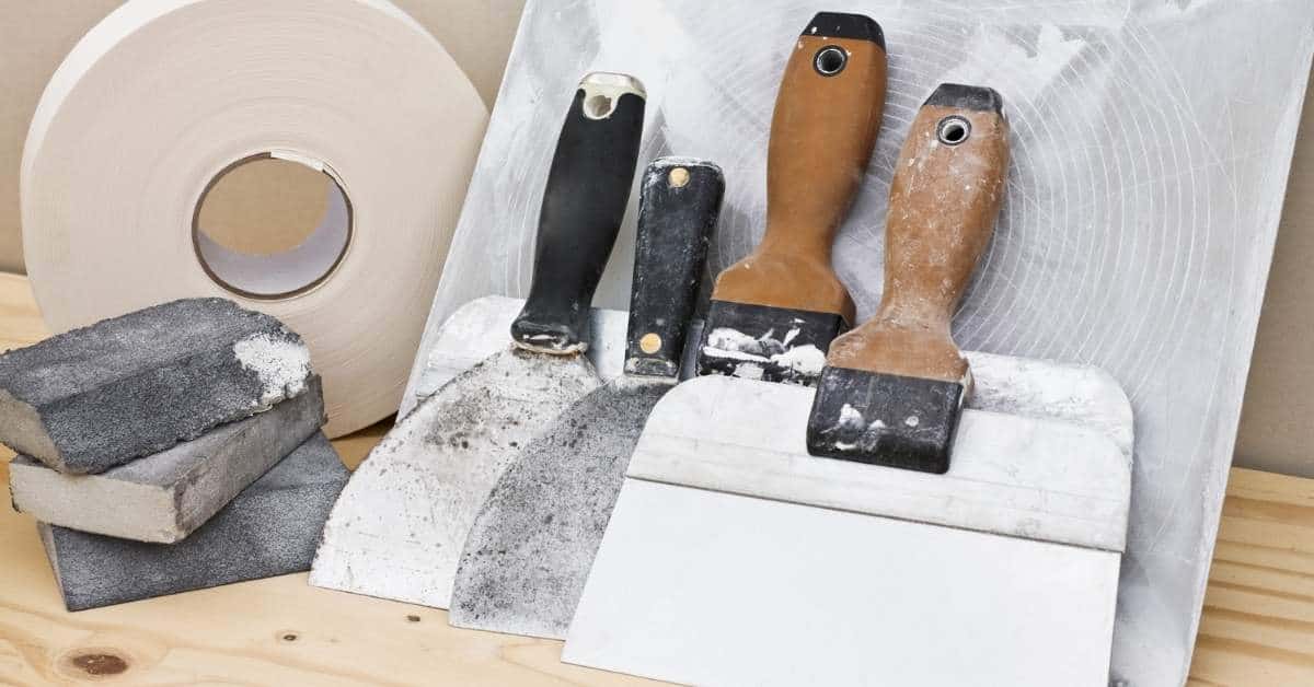 Four putty knives, sanding sponges, drywall tape roll on wood