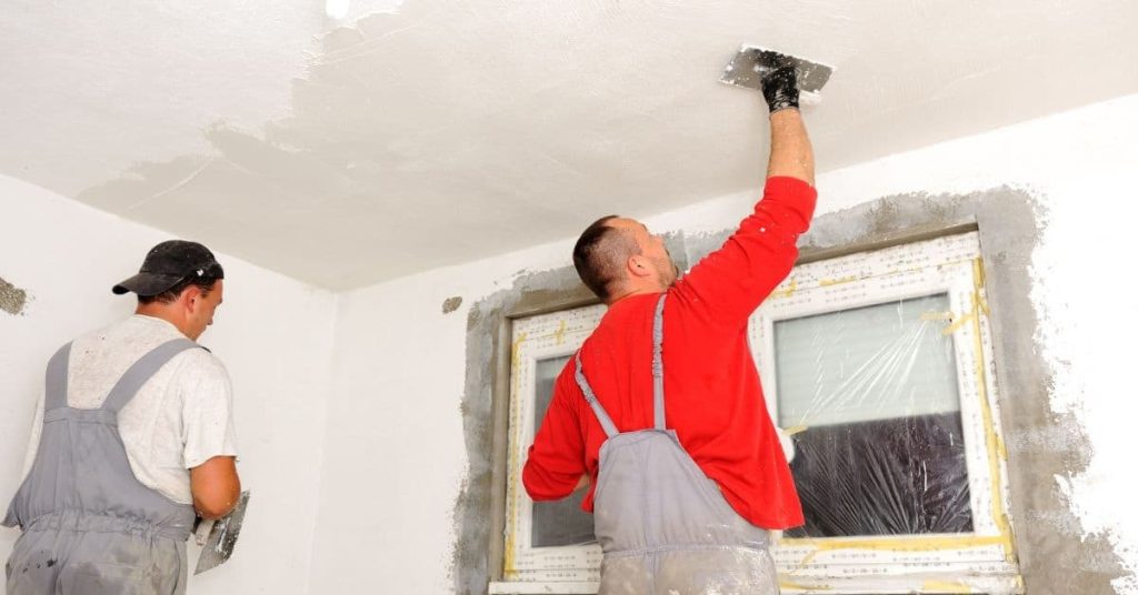 Two men applying plaster to a ceiling and wall near a window
