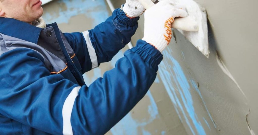 Worker applying stucco to exterior wall wearing gloves and jacket