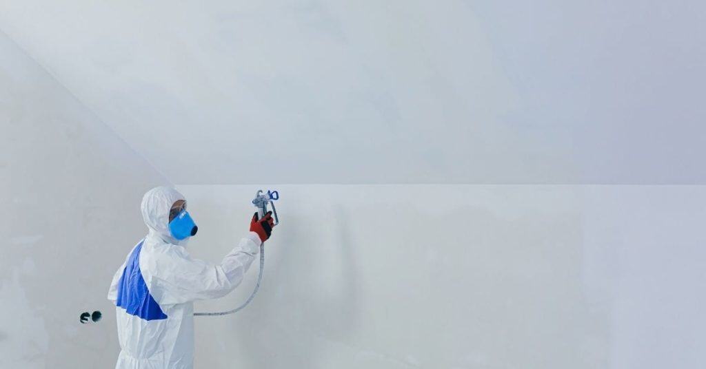Worker in protective suit spray painting white plaster wall