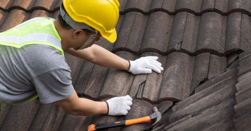 Worker inspecting broken roof tiles on sloped roof wearing yellow helmet