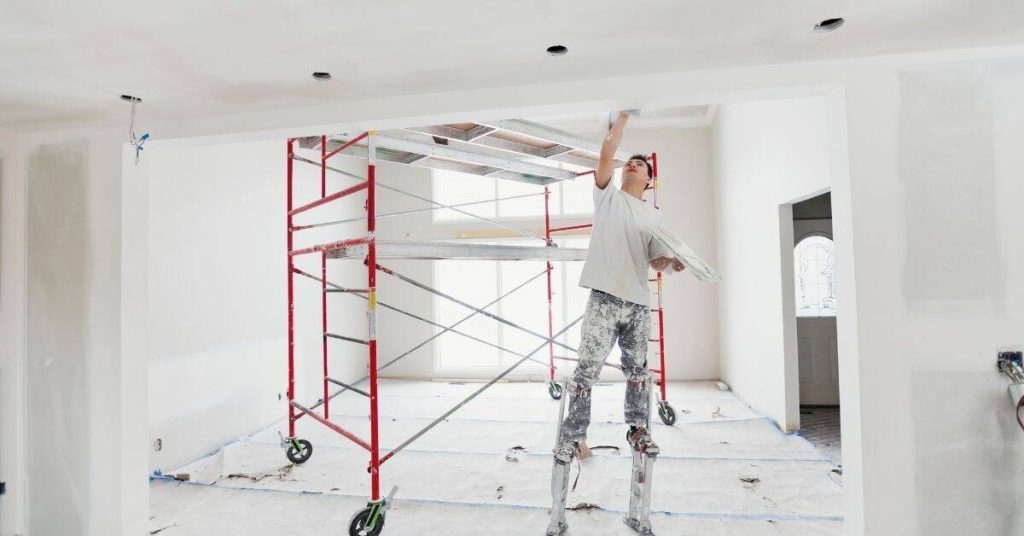 Worker on stilts applying plaster to ceiling near scaffolding