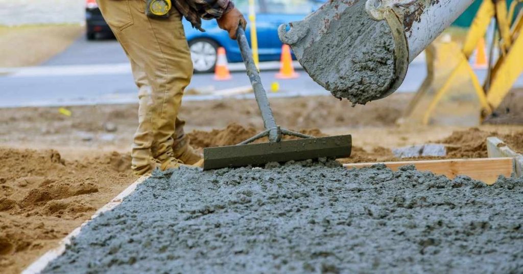 Worker smoothing wet concrete with rake near pouring excavator