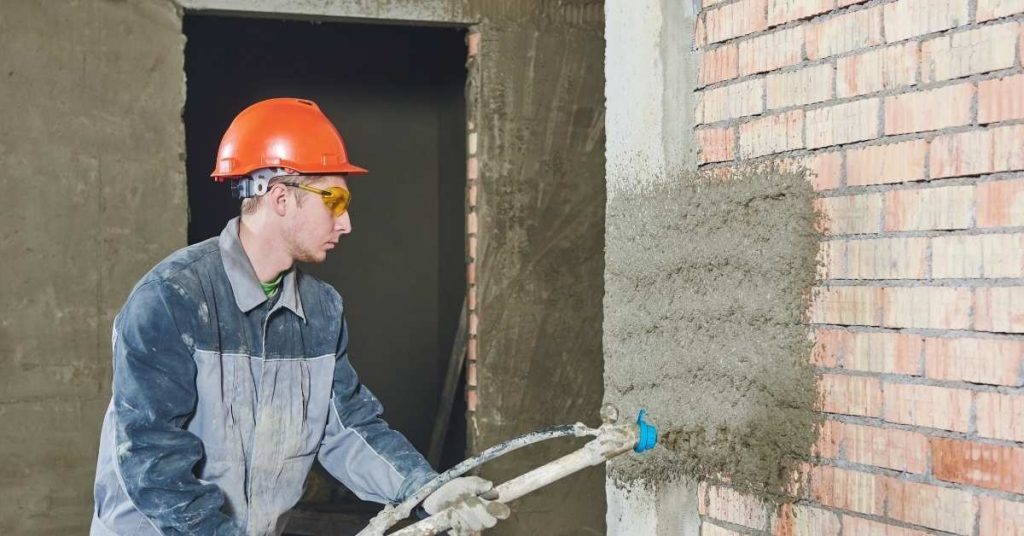 Worker spraying plaster over brick wall indoors