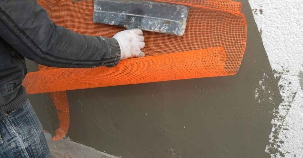 Person applying orange mesh over wet brown stucco base coat