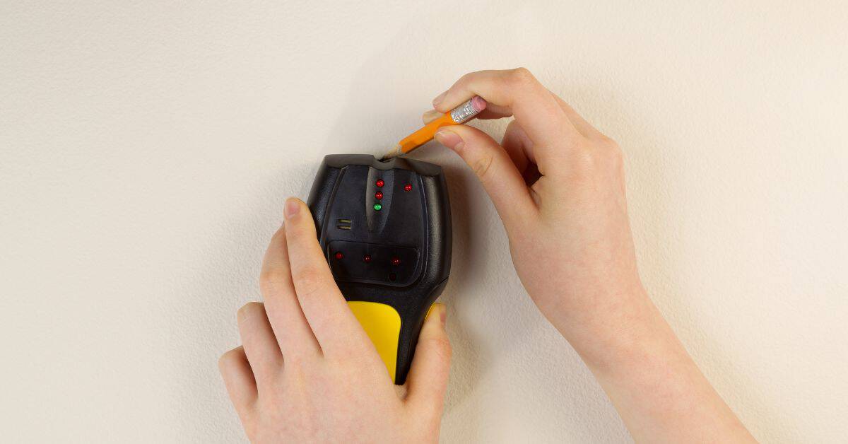 Hands holding stud finder and pencil marking beige wall