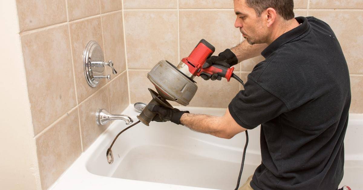 Man using electric drain cleaner in bathtub with tiled walls