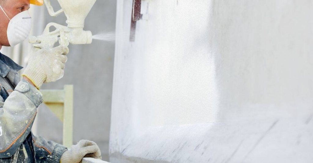 Worker spraying white coating onto stucco wall with spray gun