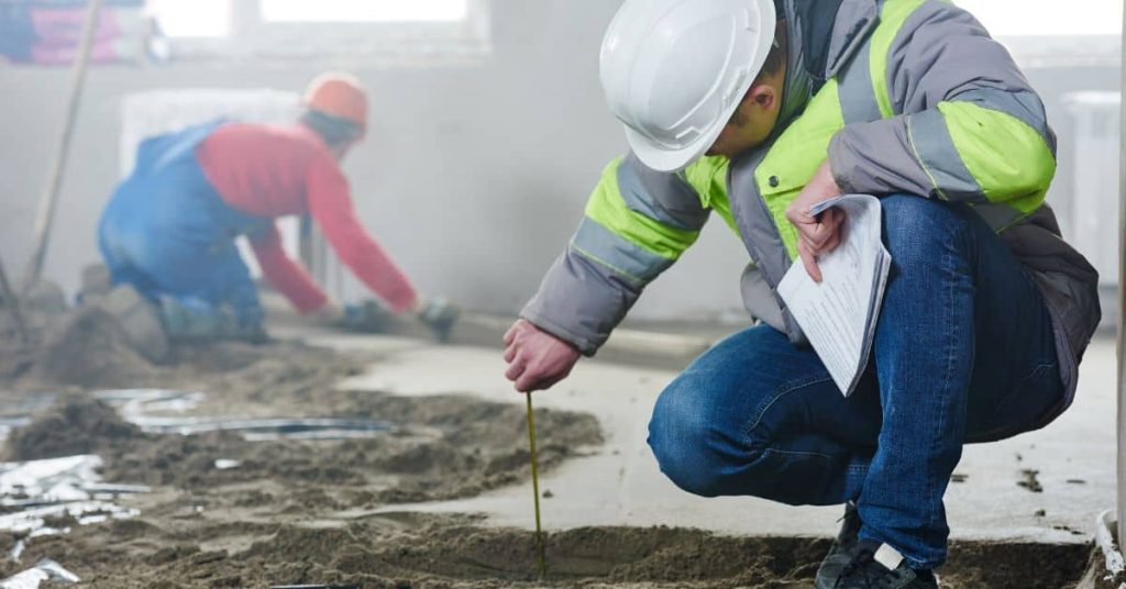Worker measuring a muddy concrete floor with another worker in the background